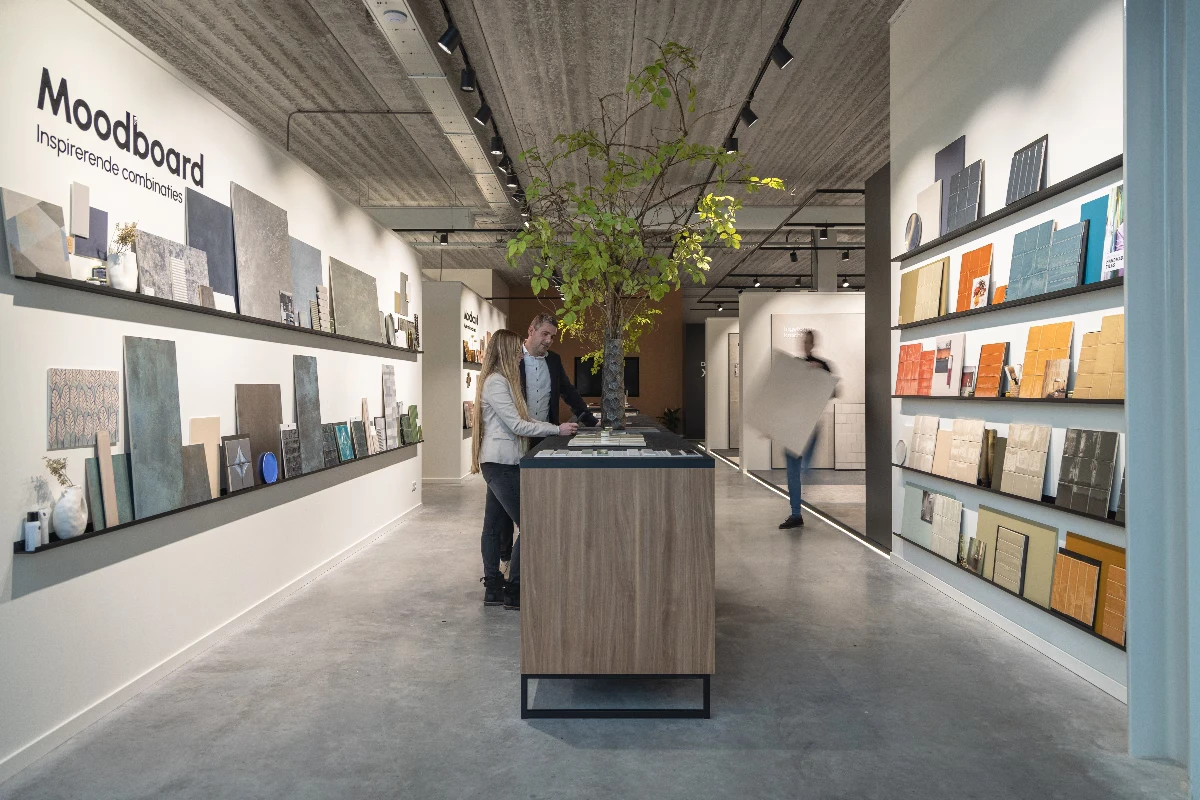 Impressie van een tegel showroom, voorzien van displays met samples van tegels en accenttegels zien. De tegels variëren in kleur, formaat en textuur. In het midden zien we een toonbank waar de tegels uitgebreid bekeken kunnen worden en een moodboard kan worden gemaakt.
