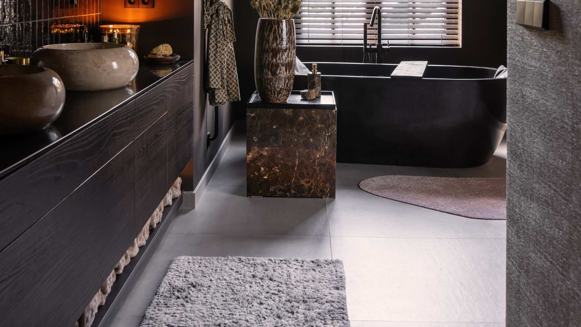 Warme, intieme badkamer met een vloer van grote grijze tegels. Bij het badkamermeubel zien we een wand van glanzende zwarte tegeltjes, en een wastafelblad in glanzend donkerbruin.