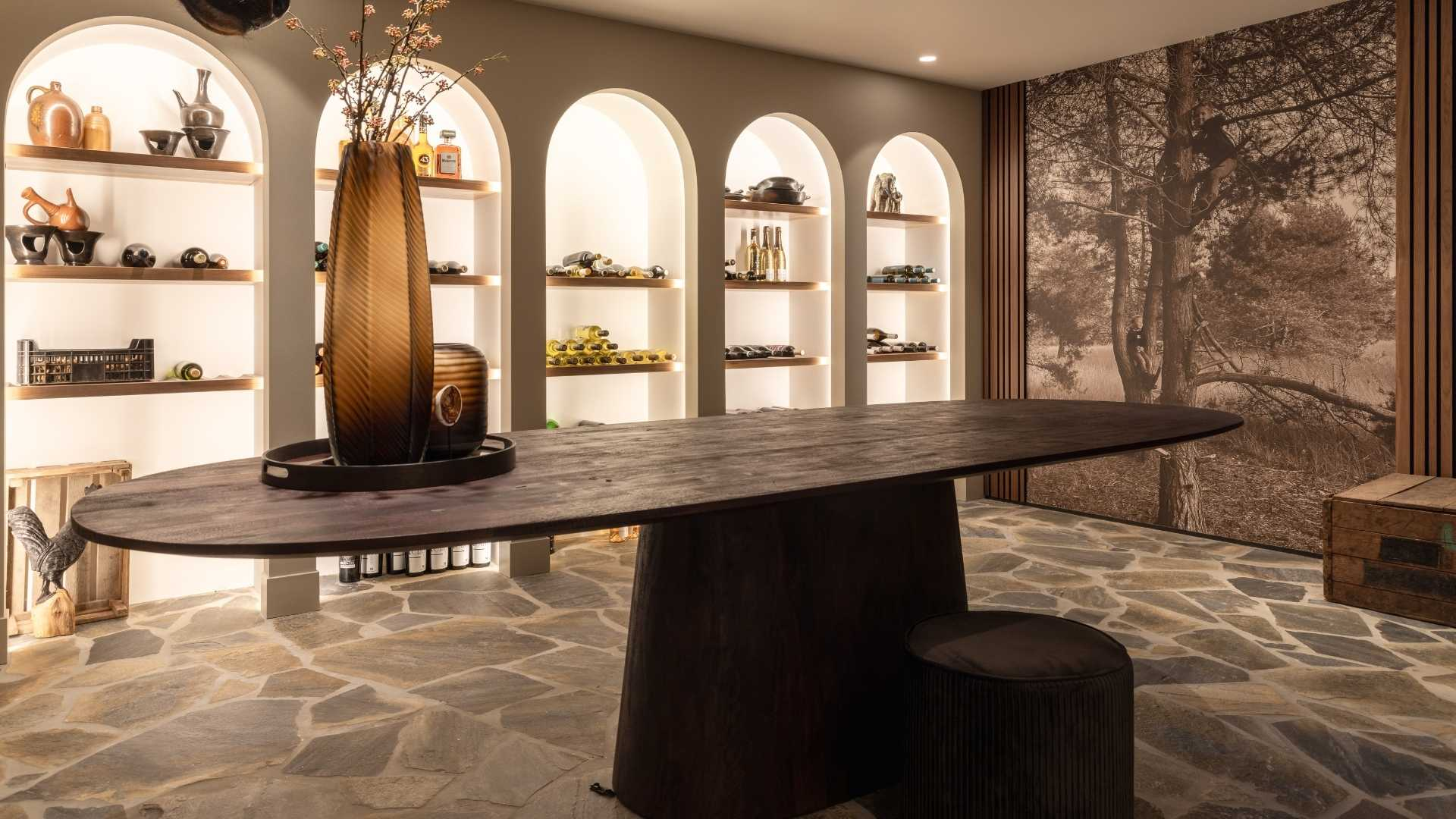 Impressie van een landelijk ingerichte wijnkelder, ingericht met flagstone tegels, zwart eiken ovale tafel en verlichte togen met wijnopslag.
