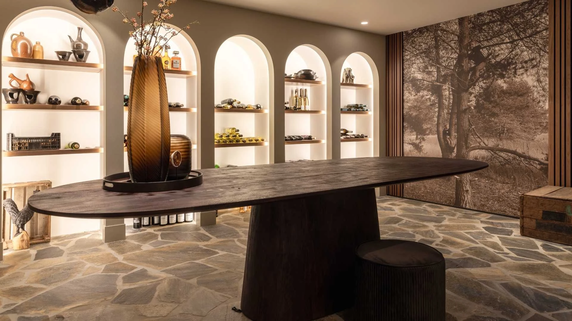Impressie van een landelijk ingerichte wijnkelder, ingericht met flagstone tegels, zwart eiken ovale tafel en verlichte togen met wijnopslag.