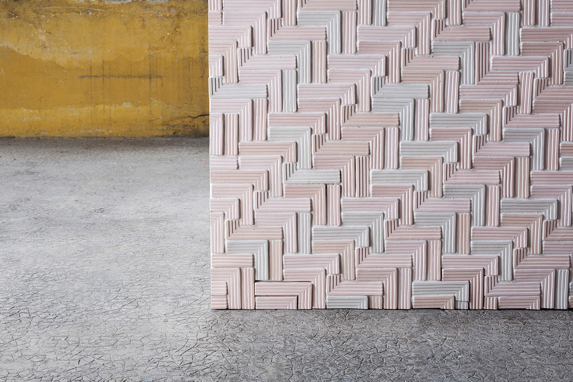 Een close-up van een wand met tegels in een patroon van pasteltinten en een textuur die lijkt op geweven stof, tegen een ruwe betonnen achtergrond.