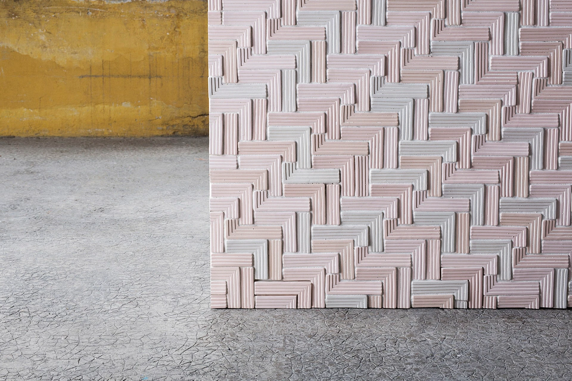 Een close-up van een wand met tegels in een patroon van pasteltinten en een textuur die lijkt op geweven stof, tegen een ruwe betonnen achtergrond.